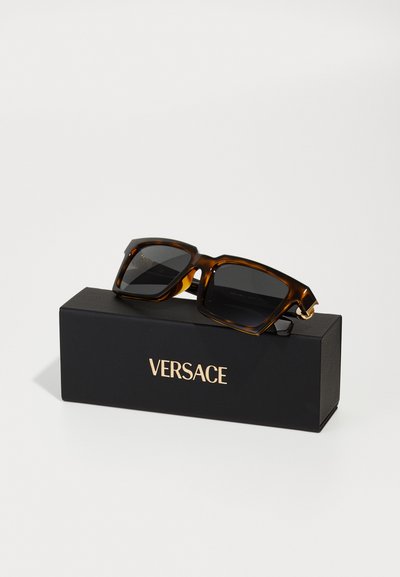 Brown tortoiseshell sunglasses on a black box, featuring dark lenses and gold accents. The box is labeled with "VERSACE" in gold lettering.