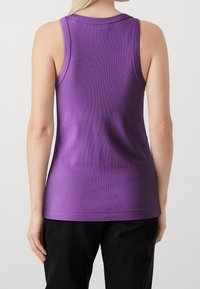 Kobieta w fioletowej, bezrękawnikowej, żebrowanej bluzce i czarnych spodniach, pokazana od tyłu na tle jasnego, jednolitego tła.