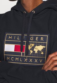 Námořnická mikina s kapucí s velkou vyšívanou nášivkou zlaté světové mapy, červenými a bílými akcenty a zlatým písmem. Šňůrky s kovovými koncovkami.