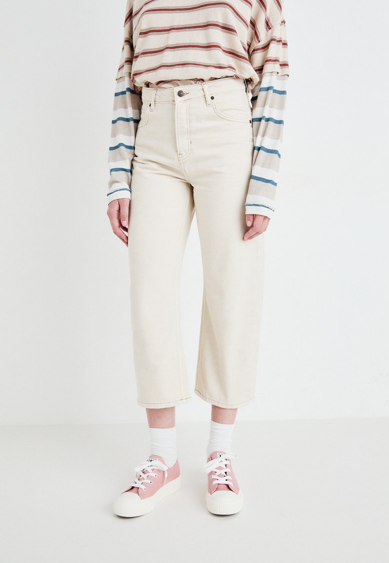 Beige wijde jeans met een hoge taille, gecombineerd met een gestreept shirt met lange mouwen in crèmekleur, blauw en rood, en roze casual sneakers.