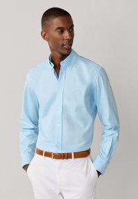 Homme portant une chemise bleu clair à boutons et un pantalon blanc avec ceinture marron, mains dans les poches, regardant sur le côté devant un fond uni.
