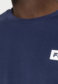 Camiseta de algodón azul marino con cuello redondo y mangas cortas, que presenta una etiqueta de logo blanca en el pecho. Textura suave, diseño minimalista.