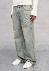 Jeans de mezclilla de corte holgado en azul claro con un acabado desgastado. Presenta un diseño de cinco bolsillos y costuras visibles. Combinados con zapatillas blancas.