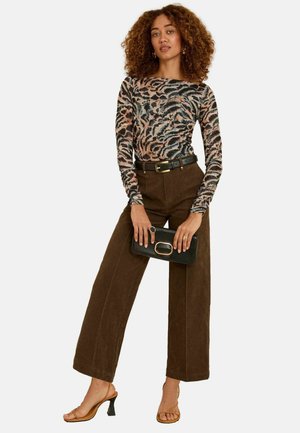 Frau mit lockigem Haar, die ein gemustertes Langarmoberteil, braune weite Hosen mit einem schwarzen Gürtel, beige Absatzschuhe trägt und eine schwarze Clutch hält.