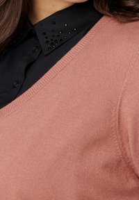 Maglione rosa con scollo a V, abbinato a una camicia nera con colletto borchiato. La trama a maglia è visibile sulla superficie del maglione.
