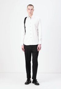 Biely košeľový top s zelenými akcentmi, spárovaný s čiernymi nohavicami a čiernymi topánkami. Model stojí vzpriamene s čiernou ramennou taškou.
