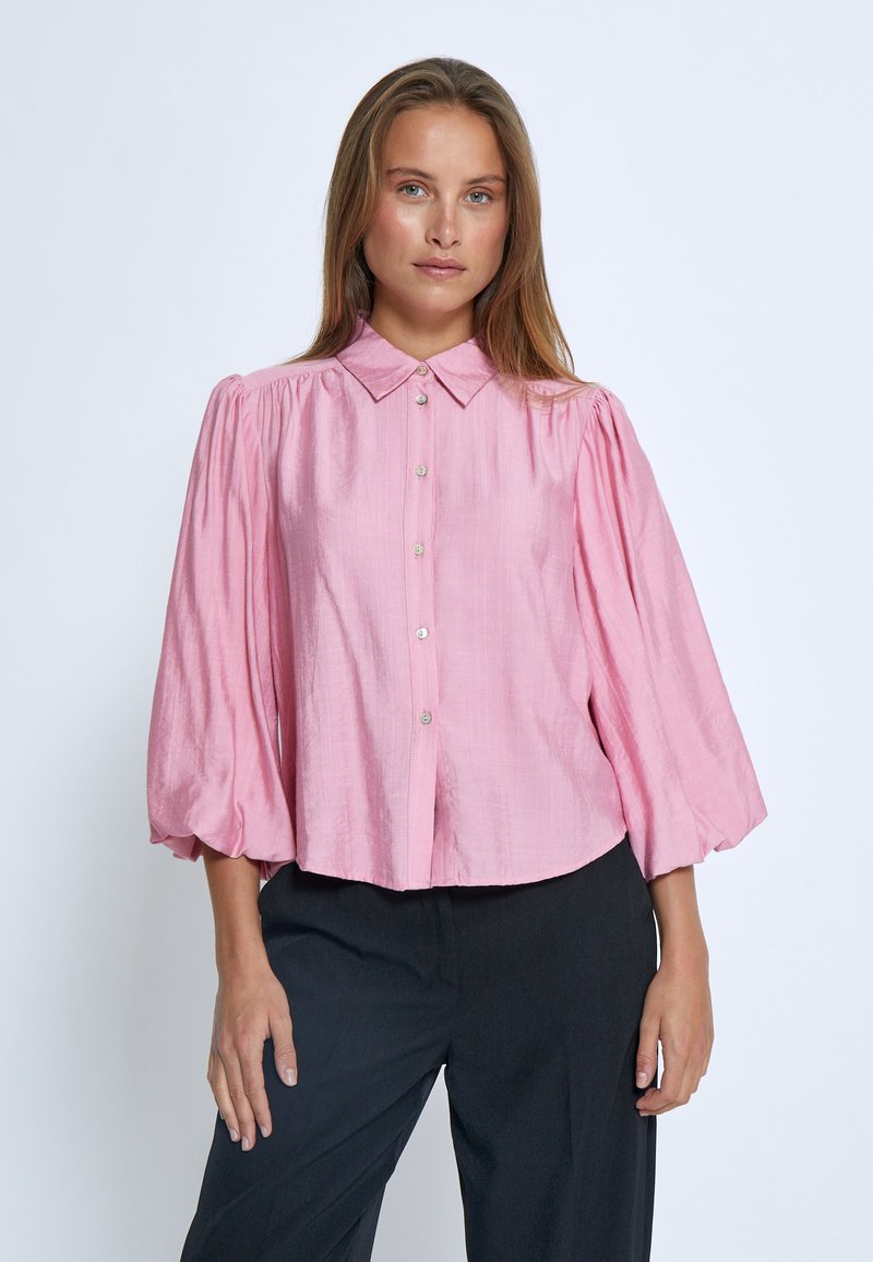 Blusa rosa con colletto e maniche a palloncino, chiusura con bottoni e un tessuto leggermente testurizzato. Indossata con pantaloni neri per creare un contrasto.