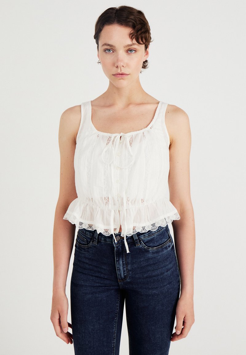 Witte cropped tanktop met kantafwerking, met een strik aan de voorkant en knoopsluiting. Gecombineerd met donkerblauwe skinny jeans.