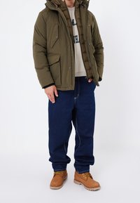 Chaqueta acolchada verde oliva con dos bolsillos delanteros, combinada con vaqueros azules y botas marrones con cordones dorados. Textura suave y diseño aislante.