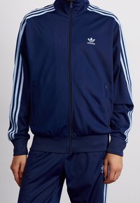 Marinblå träningsjacka i mjuk polyester, med hög krage, dragkedja och ljusblå tre-randsdetaljer på ärmarna. Logotyp på bröstet.