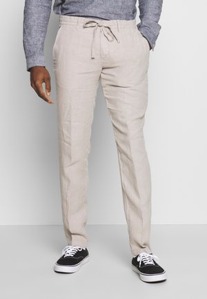 Man met beige broek met trekkoord, zwart-witte sneakers en een grijs shirt met lange mouwen, staand met één hand in de zak.