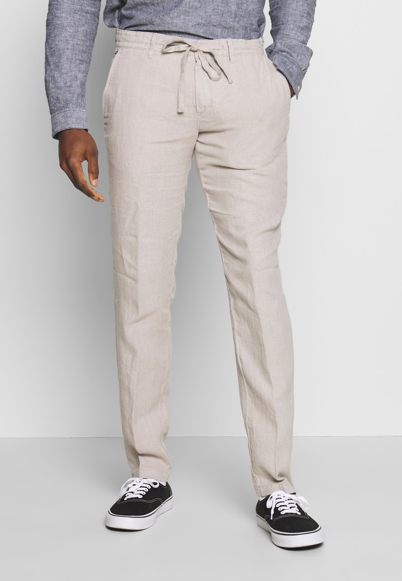 Homme portant un pantalon beige à cordon, des baskets noires et blanches et une chemise grise à manches longues, debout avec une main dans la poche.