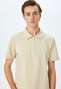 Beige polo-shirt med korte ærmer, krave og halvmåneslås. Fremstillet af glat stof, har den rene linjer og et minimalistisk design.