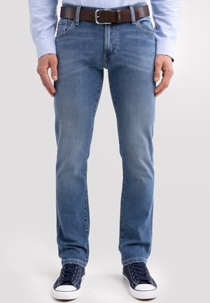 Vyras dėvi šviesiai mėlynas džinsus, rudą odinį diržą, tamsiai mėlynas sportbačius ir šviesiai mėlyną ilgomis rankovėmis marškinį, stovintis prieš paprastą foną.