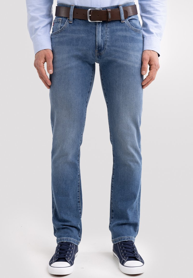 Vyras dėvi šviesiai mėlynas džinsus, rudą odinį diržą, tamsiai mėlynas sportbačius ir šviesiai mėlyną ilgomis rankovėmis marškinį, stovintis prieš paprastą foną.