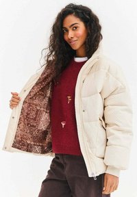 Krémová péřová bunda s texturovaným povrchem, zapínáním na patentky a vzorovanou hnědou vnitřní podšívkou. Kombinována s bordovým svetrem a tmavými kalhotami.