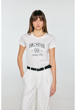 Młoda kobieta o prostych brązowych włosach, ubrana w białą koszulkę z napisem "Big Star Sailing Team" oraz białe spodnie z czarnym paskiem, stojąca na jednolitym tle.