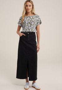 Blusa floreale verde con maniche arrotolate, abbinata a una gonna in denim nera a vita alta con uno spacco frontale, indossata con sneakers grigie.