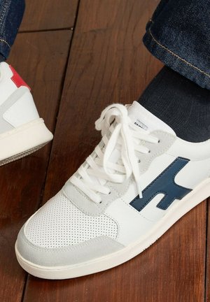 Witte sneaker met blauw logo en grijze suède accenten, gedragen met donkere sokken en jeans op een houten vloer.