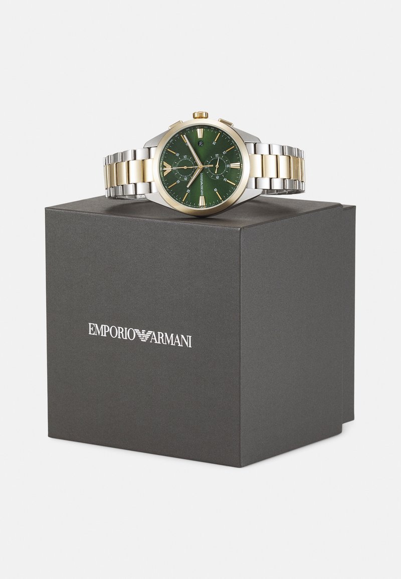 Eine goldene und silberne Edelstahl-Uhr mit grünem Ziffernblatt, leuchtenden Zeigern, zwei Hilfszifferblättern und einem Logo. Präsentiert in einer dunklen Box.