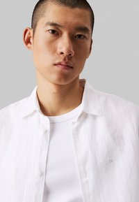Camicia bianca in lino con bottoni, indossata sopra un top a canotta bianco a coste, caratterizzata da un logo discreto e un colletto, con una texture liscia e una vestibilità comoda.