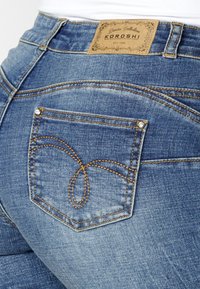 Jean en denim bleu avec une étiquette de ceinture visible, un design unique de surpiqûres orange sur la poche arrière, et un petit rivet décoratif.