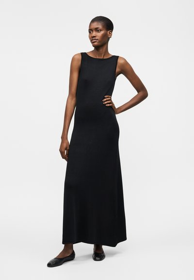 Frau steht mit der Hand in der Hüfte, trägt ein ärmelloses schwarzes Maxikleid und schwarze flache Schuhe vor weißem Hintergrund.