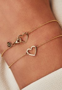 Bracelet en chaîne dorée présentant deux charms en forme de cœur ouvert, un fermoir à homard et une petite étiquette logotypée. Texture lisse avec finition polie.