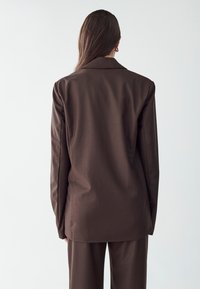 Hnedý blazer s hladkou textúrou, priliehavým strihom a zárezy na golieri, zobrazený zozadu. Koordinované nohavice sú tiež viditeľné.