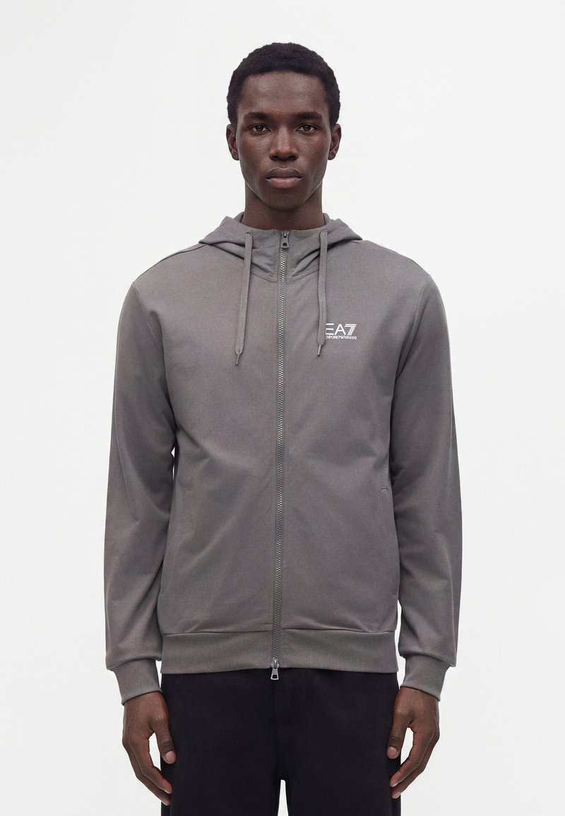 Jeune homme portant un sweat à capuche gris zippé avec le logo EA7 Emporio Armani sur le côté gauche de la poitrine, debout devant un fond blanc uni.