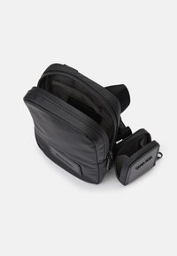 Geantă crossbody din nylon negru, cu o textură netedă, închidere cu fermoar și un pachet mai mic detasabil. Are un patch frontal și o curea reglabilă.