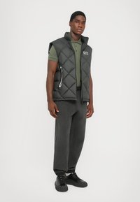 Vest negru matlasat, fără mâneci, cu guler înalt și buzunare cu fermoar, asortat cu un polo verde, pantaloni gri și adidași negri.