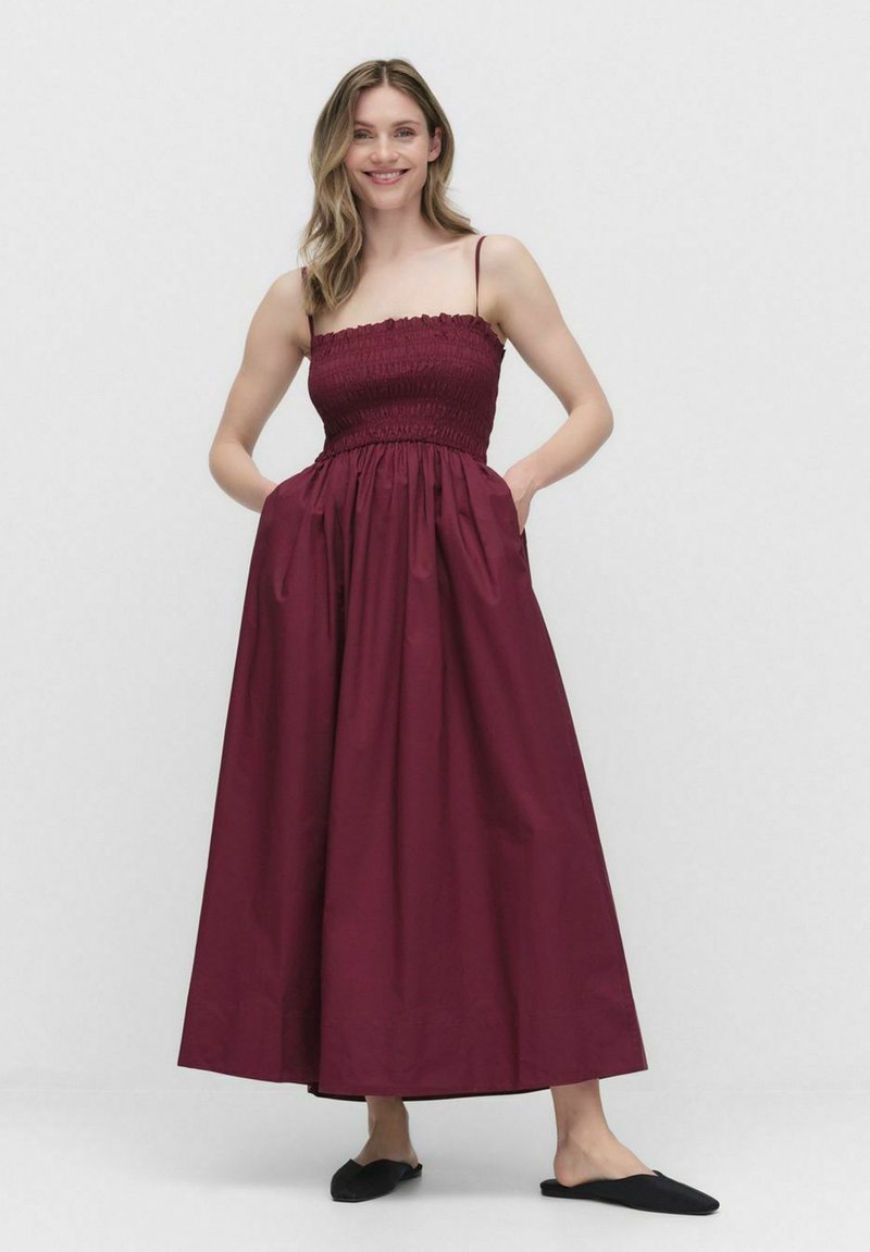 Burgundy ärmlös maxiklänning med smockad bodice, sidofickor och en flödande kjol; tillverkad av lättviktig tyg.