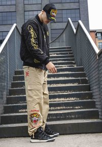 Svart bomberjacka med gul broderi, beige cargobyxor med tigeremblem och svarta sneakers med silverdetaljer, poserar på trappor.