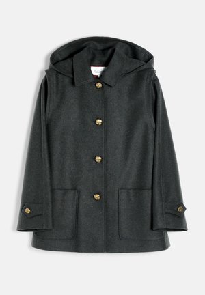 Jeune femme portant une casquette noire, une veste gris foncé, un pull à col roulé rouge, un pantalon bordeaux et des bottes noires montantes, debout les mains dans les poches.