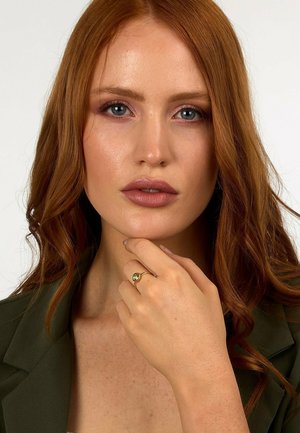 Goldener Ring mit einem grünen Stein, getragen an einer Hand mit heller Haut. Das Modell hat rote Haare, ein neutrales Make-up und trägt ein dunkelgrünes Sakko.