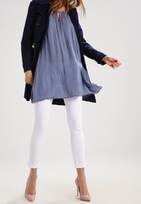 Cappotto trench navy su una blusa leggera e fluida azzurra, abbinato a jeans skinny bianchi e tacchi alti rosa pallido. Tessuto morbido e vestibilità rilassata.