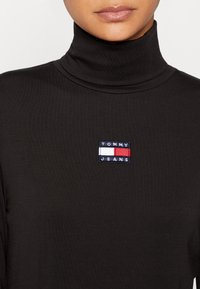 Kvinna i svart höghalset tröja med liten Tommy Jeans-logotyp på bröstet, endast läppar och nacke synliga.