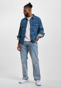 Blå denimjakke med to brystlommer, brukt over en hvit T-skjorte. Lyseblå slitte jeans og hvite sneakers med blå detaljer.