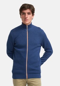 Blå zip-sweater med høy krage, med et teksturert mønster og oransje glidelåsdetaljer. Ribbestrikkede mansjetter og hem fullfører designet.