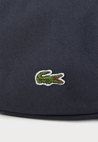 Mörk marineblått tekstil med en grön krokodil-logo broderad med vita detaljer, satt mot en teksturert bakgrunn.