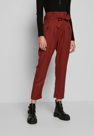Mujer con pantalones de talle alto color rojizo óxido con cinturón atado y botones negros, combinados con botines negros y un top de manga larga negro.