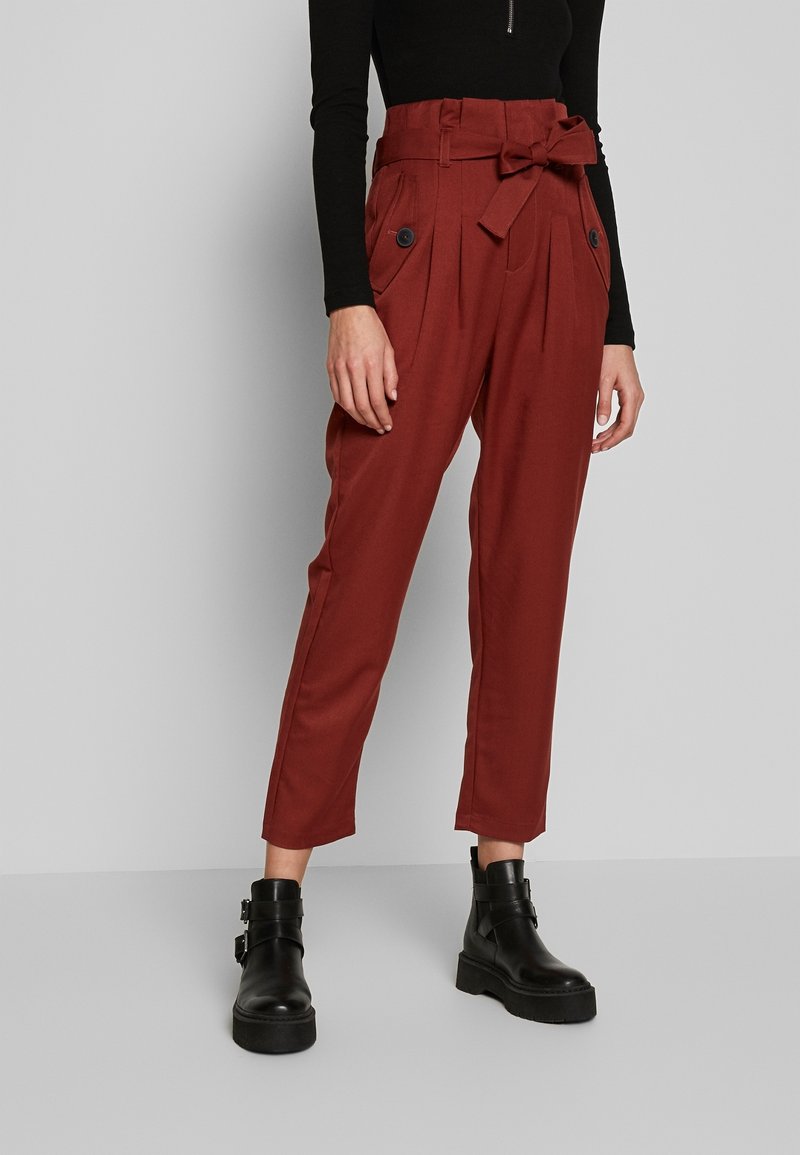 Femme portant un pantalon taille haute rouge rouille avec une ceinture nouée et des boutons noirs, associé à des bottines noires et un haut noir à manches longues.