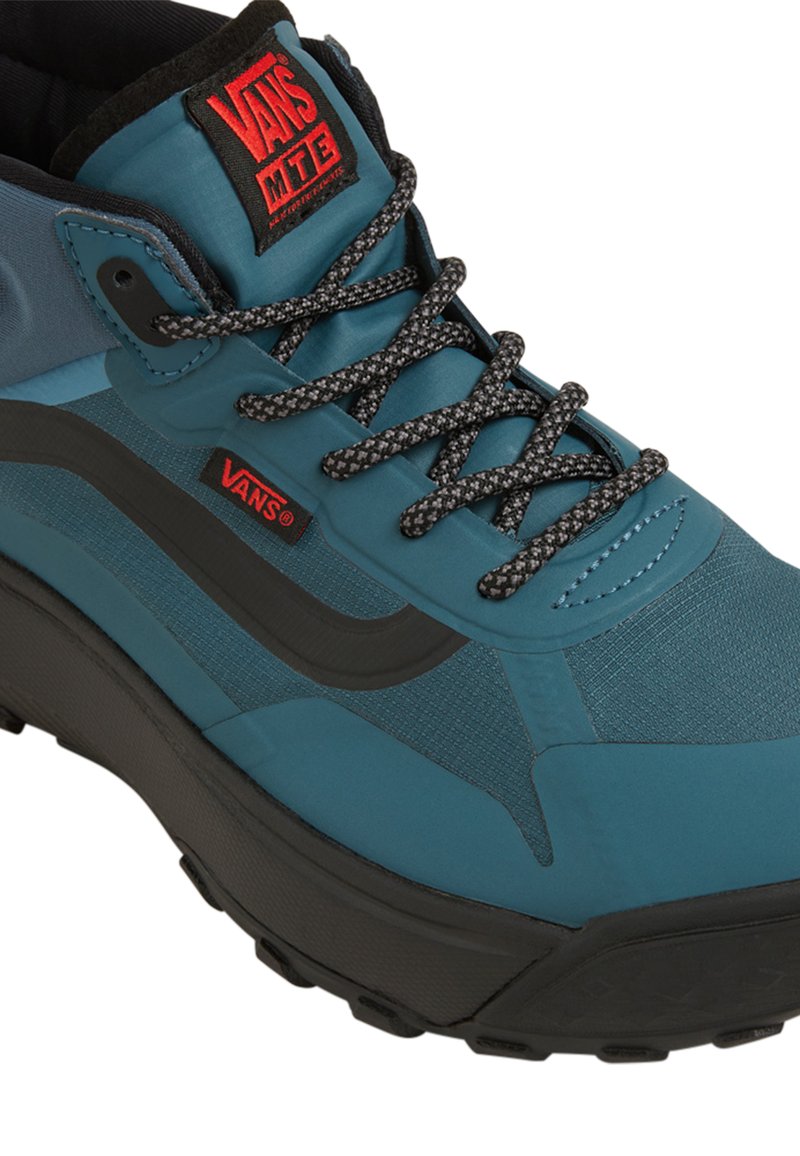 Zapatilla alta color turquesa con tejido texturizado, acentos negros, cordones grises y un parche con el logotipo de Vans en la lengüeta. Suela de goma para mayor agarre.