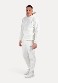 Felpa bianca con cappuccio e pantaloni abbinati, in morbido materiale felpato, tasca frontale sulla felpa, branding minimale, indossata con sneakers bianche.