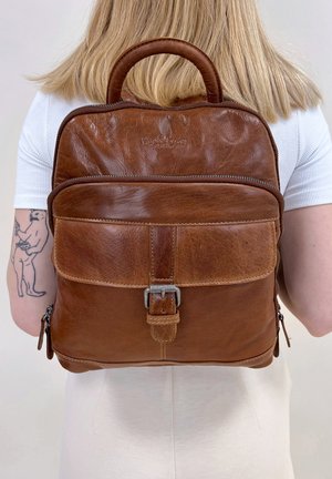 Brown leather backpack with front buckle pocket worn by person with blonde hair and tattoo on left arm, against plain background.