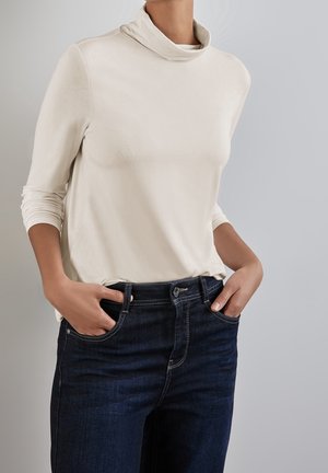 Personne portant un t-shirt à col roulé crème à manches longues et un jean bleu foncé, les mains dans les poches, sur un fond uni.