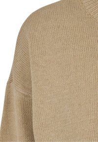 Pull en tricot beige, avec une surface texturée, un col en V et des manches larges avec un motif côtelé le long du col et des épaules.