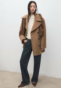 Cappotto in lana color cammello con colletto oversize e chiusura con doppiopetto a bottoni, abbinato a maglione crema e pantaloni in denim scuro, completato da scarpe marroni con punta appuntita.