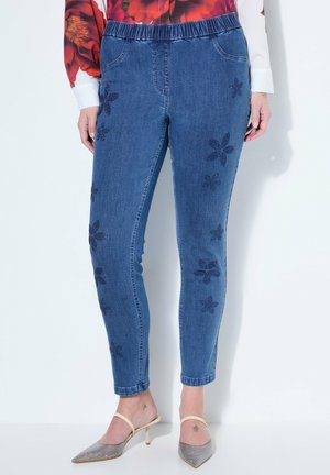 Frau trägt blaue Denim-Leggings mit elastischem Bund und dunklen Blumenmustern, eine rote Bluse mit Blumenmuster und spitze Pumps mit Knöchelriemen.